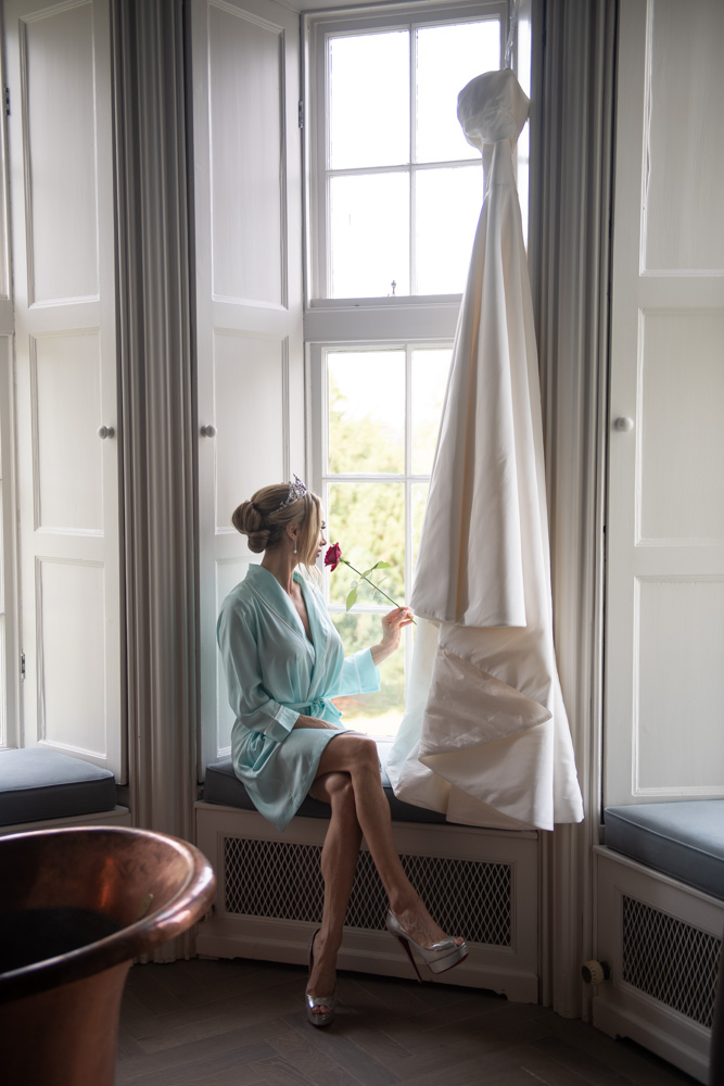 bride sitting in window beside hanging wedding dress