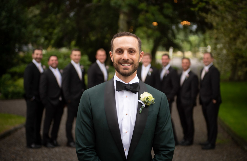 Groom in green suit in foreground and his groomsmen in the background all looking at camera