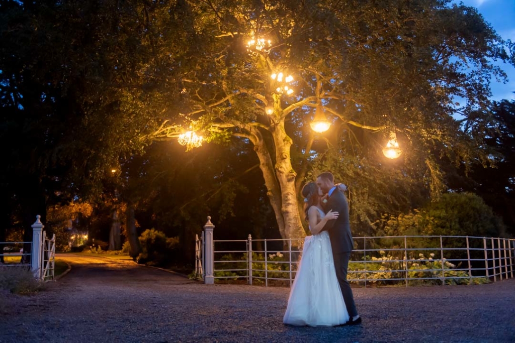 Bride and groom outside at night time kissing under the tree full of lit chandeliers at Ballymagarvey