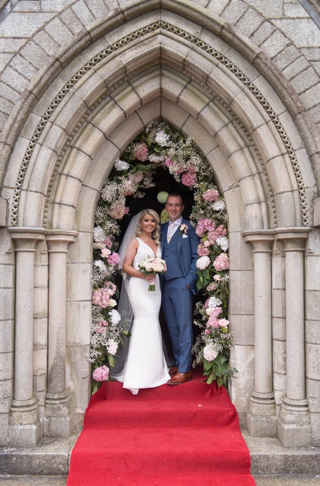 Bride and groom standing in the Church door with a white and pink flower arch