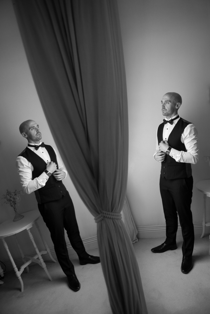 Black and white of groom standing looking out the window with a reflection of him in the mirror