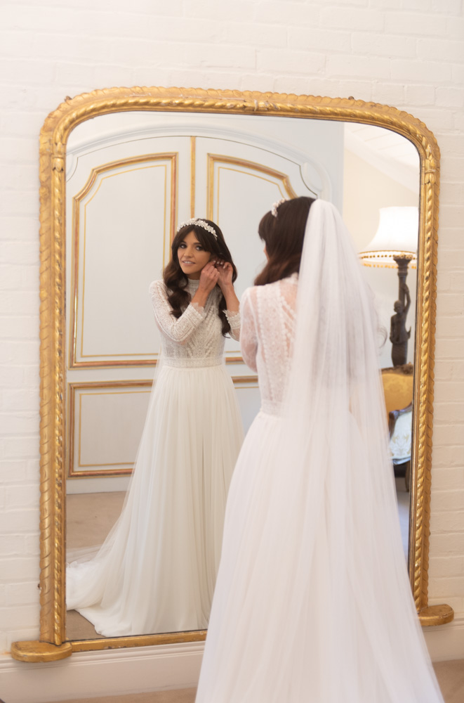 Bride in her wedding dress putting in her earring while looking in the mirror