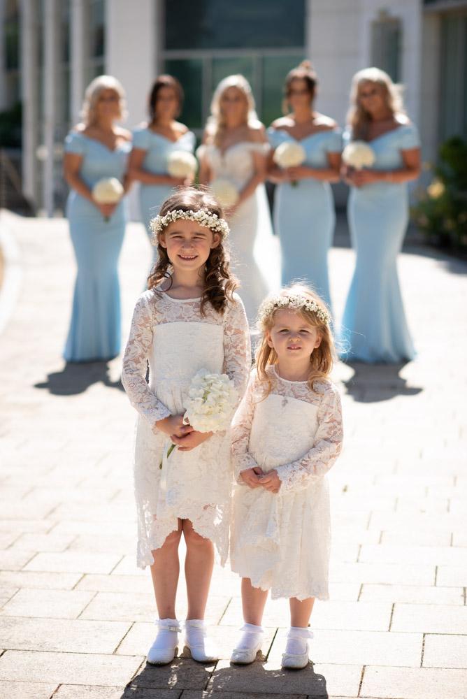 Two flower girls in white dresses standing in the foreground looking at camera and the Bride and Bridesmaids in the background