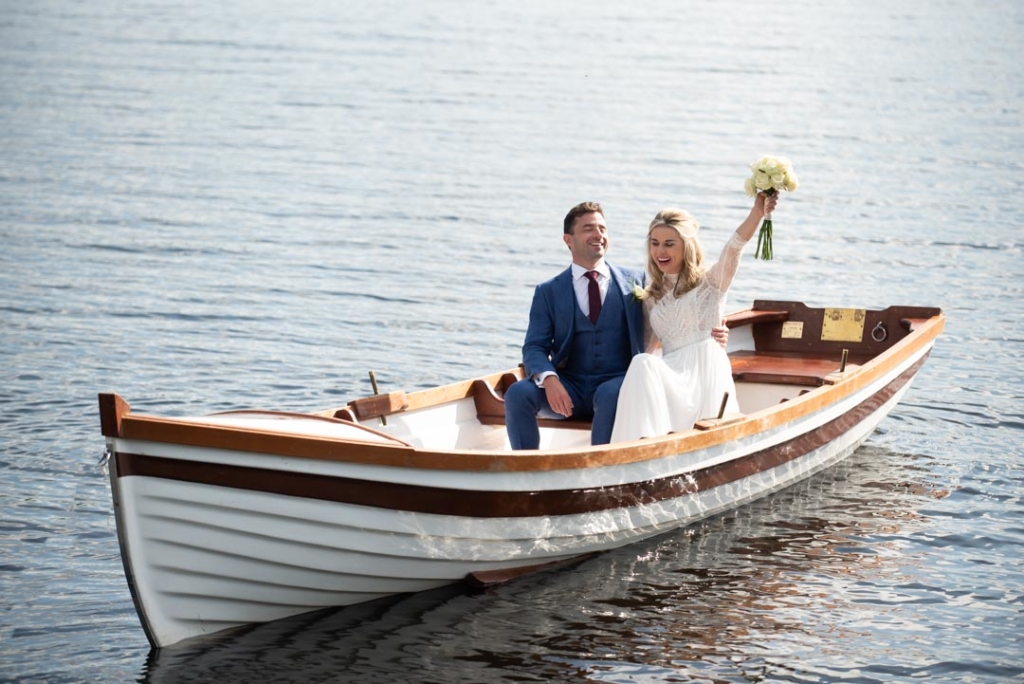 Bride with her hand and flowers in the air and groom sitting beside her in a boat on the lake at Virginia Park Lodge